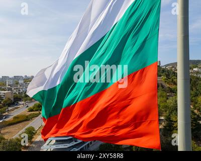 La bandiera bulgara ondeggia orgogliosamente sopra una città costiera, con il vasto mare e il verde paesaggio sullo sfondo. Un simbolo di orgoglio nazionale e.. Foto Stock