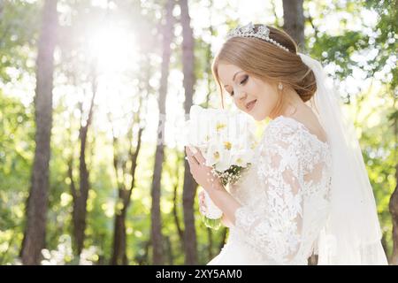 Ritratto di una bella sposa in un grande e lussureggiante abito bianco con un bouquet di fiori in mano tra i caldi raggi del sole in una foresta verde Foto Stock