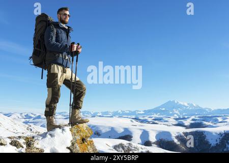 Un viaggiatore hipster con la barba che indossa occhiali da sole in natura. Un uomo che cammina in montagna con uno zaino e bastoni da passeggio scandinavi nel bac Foto Stock