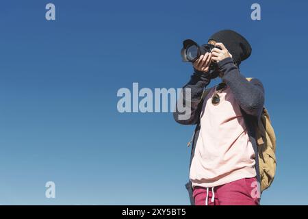 Ritratto di una ragazza hippy elegante con un cappello e uno zaino che la fotografa con una fotocamera reflex digitale all'aperto contro un cielo blu in una zona soleggiata Foto Stock