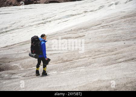 Un alpinista con uno zaino cammina in carrozzina, si erge su un ghiacciaio polveroso con scarpe da ginnastica tra le fessure delle montagne Foto Stock