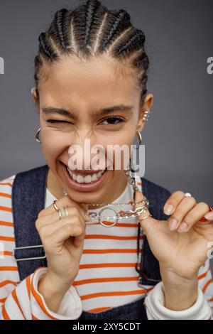 Ritratto verticale di una giovane donna etnica con acconciature di cordoglio che posa con atteggiamento in studio e che si fa ammiccante alla macchina fotografica giocando con gioielli Foto Stock