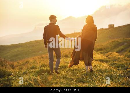 Una giovane coppia. Il ragazzo conduce una ragazza riccia avvolta in un plaid con un bouquet di fiori al tramonto. Il concetto di fiducia e famiglia giovane felice Foto Stock