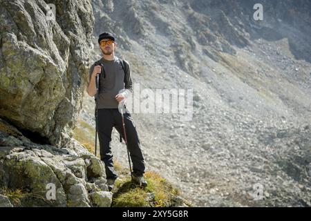 Porter di un elegante viaggiatore hipster con una barba e uno zaino in occhiali da sole e un berretto con pali da trekking si erge su una roccia sullo sfondo di Foto Stock