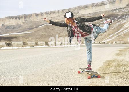 Elegante ragazza Happy Young con berretto e tuta in denim, in equilibrio tra una strada di montagna e un longboard Foto Stock