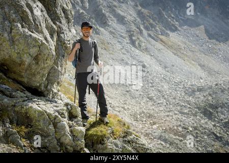 Porter di un elegante viaggiatore hipster con una barba e uno zaino in occhiali da sole e un berretto con pali da trekking si erge su una roccia sullo sfondo di Foto Stock