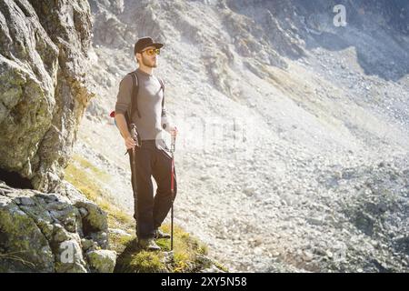 Porter di un elegante viaggiatore hipster con una barba e uno zaino in occhiali da sole e un berretto con pali da trekking si erge su una roccia sullo sfondo di Foto Stock