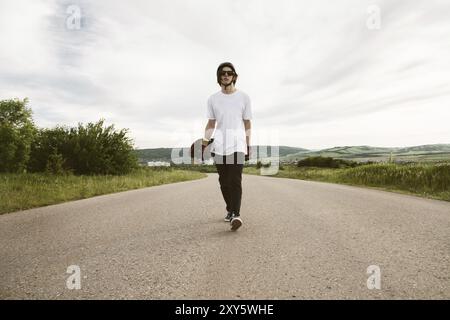 Un ragazzo con un longboard in mano con un casco e occhiali da sole cammina lungo la strada di campagna asfaltata in un viaggio Foto Stock