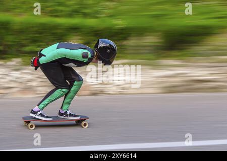 Un giovane con casco integrale e una tuta in pelle in uno speciale rack, cavalca un longboard sul tramonto afsaltu sulle montagne sullo sfondo e sul bellissimo sk Foto Stock