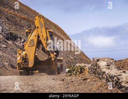 Escavatore giallo che viaggia lungo una strada polverosa e un uomo aggrappato al suo fianco Foto Stock