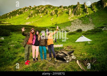 Un gruppo di campeggiatori ritratti si trova vicino a una tenda bianca in un lussureggiante e verdeggiante scenario montano. I turisti sorridenti ondeggiano allegramente. Scogliere rocciose e vegetazione vibrante, che creano un'atmosfera mozzafiato e avventurosa. Foto Stock