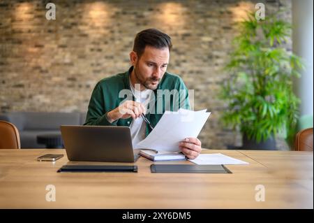 Un serio contabile maschile che tiene gli occhiali e analizza i report finanziari con un laptop sulla scrivania in ufficio Foto Stock