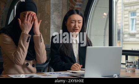 Due donne d'affari multirazziali donna asiatica coreana ragazza musulmana hijab islamico donna d'affari ufficio donne d'affari che guardano schermo portatile terribile Foto Stock