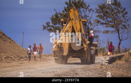 Escavatore giallo che viaggia lungo una strada polverosa e un uomo aggrappato al suo fianco Foto Stock