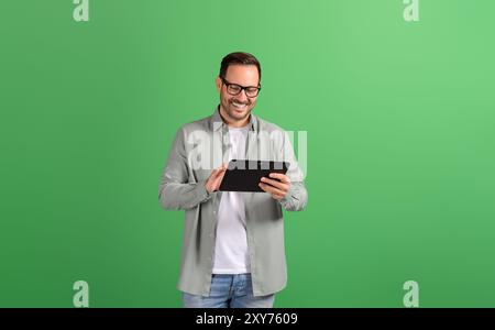 Un giovane uomo d'affari sorridente con gli occhiali che legge e-mail su un tablet digitale su sfondo verde Foto Stock