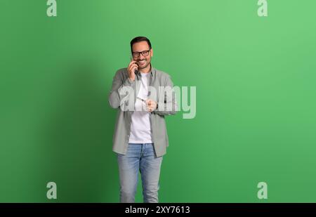 Ritratto di un bel freelance che tiene in mano la carta di credito e parla sullo smartphone su sfondo verde Foto Stock