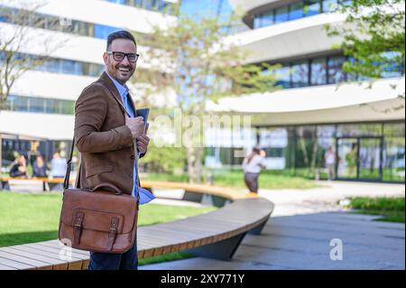 Vista laterale ritratto di un giovane uomo d'affari con laptop e borsa in piedi sul vialetto contro gli edifici Foto Stock