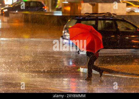 Donna che attraversa la strada in un centro urbano, tenendo in mano un ombrello colorato durante un forte temporale notturno. Foto Stock