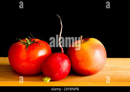 Splendida natura morta di pomodoro, mela e ravanello su un tavolo di legno su sfondo nero con luce naturale Foto Stock