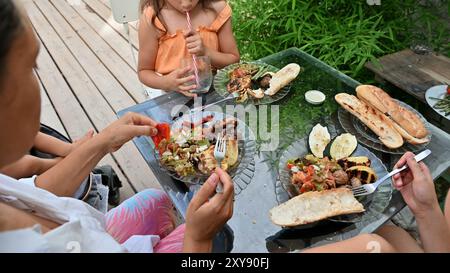 Una famiglia si riunisce per un pasto informale all'aperto, con verdure e pane alla griglia, adagiato su un tavolo di vetro in mezzo a una vegetazione lussureggiante. Foto Stock