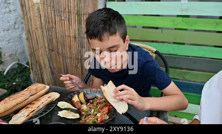 Un bambino si diverte a gustare un pasto delizioso con pane e verdure all'aperto in un vivace e colorato patio. Foto Stock