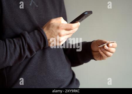 Le mani maschili hanno un telefono cellulare e una carta di credito, un giovane con un maglione dark casual paga per gli acquisti, effettua online banking utilizzando un applicat Foto Stock