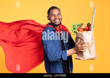 Un adulto afroamericano sorridente e che si comporta come un supereroe con un sacchetto di verdure appena raccolte, promuovendo uno stile di vita sostenibile. Giovane uomo che raccomanda nutrizione vegana in studio. Foto Stock