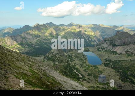 Una vista dall'alto di una catena montuosa con alte cime appuntite e un ampio lago allungato ai piedi in una soleggiata giornata estiva. Rock Parabola, Ergaki Natur Foto Stock