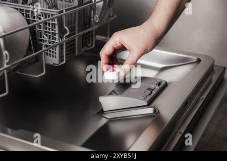 Primo piano della mano femmina inserimento di una tavoletta da lavastoviglie in una lavastoviglie automatica in acciaio inox con utensili sporchi all'interno in un moderno ambiente Foto Stock