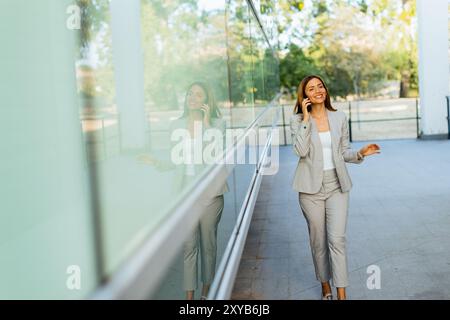 Una donna professionista cammina con facilità, sorridendo mentre si concentra sul telefono, mescolando lo stile di vita moderno con la tranquillità della natura sullo sfondo Foto Stock