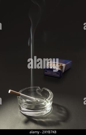 Una sigaretta di copertura sta bruciando in un posacenere, con il fumo che galleggia in aria, con un pacchetto aperto di sigarette sullo sfondo Foto Stock