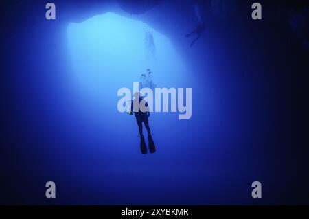 Subacquea femmina subacquea che si tuffa in una grotta galleggiante che apre un grande buco nell'ingresso del fondo marino della cavità verticale subacquea nella barriera corallina Foto Stock