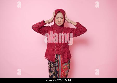 Donna asiatica stressata che indossa un moderno abito kebaya e hijab che si sente stanca e mal di testa, isolata da sfondo rosa. Foto Stock