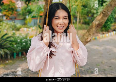 Bella giovane asiatica che indossa un abbigliamento casual sorridente alla macchina fotografica seduta e che gioca su un swing durante il weekend. Foto Stock