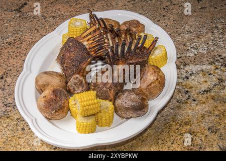 Tre rastrelliere di agnello ly in una piastra di ceramica bianca che si trova su una superficie di granito. L'incontro è circondato da patate al forno e mais bollito il giorno Foto Stock