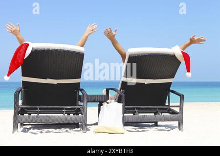 Cartolina di Natale o sfondo, coppia in lettini con cappelli di Babbo Natale in piedi su una splendida spiaggia tropicale con sabbia bianca e acqua turchese. Concetto Foto Stock