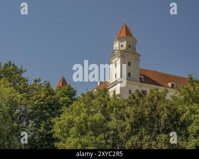 Edificio del castello con torri e tetto arancione dietro le cime degli alberi sotto un cielo blu, bratislava, slovacchia Foto Stock