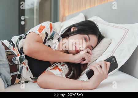La giovane donna asiatica triste sta sdraiata su un letto e guarda il suo smartphone mentre si pulisce le lacrime dopo aver pianto. Foto Stock