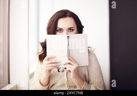 Giovane e bella donna bruna che indossa un maglione in maglia leggendo un libro che si rilassa vicino alla finestra. Concetto di relax in casa Foto Stock