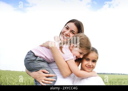 Piacere giovane madre ridere e guardare la macchina fotografica e abbracciando piccole figlie con campo verde e cielo sullo sfondo Foto Stock