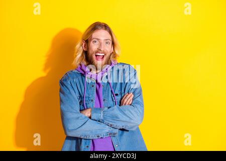 Foto di un giovane uomo simpatico con braccia piegate e spazio vuoto. Indossa una giacca in denim isolata su sfondo giallo Foto Stock