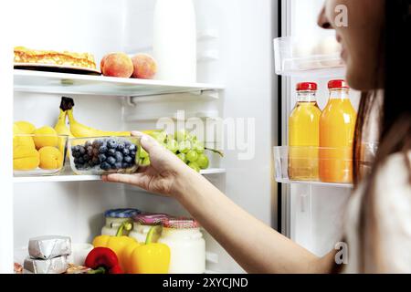 Donna che prende, afferra o preleva una scatola di mirtilli dal ripiano aperto del frigorifero o dal cassetto del frigorifero pieno di frutta, verdura, banana, pesche, Foto Stock