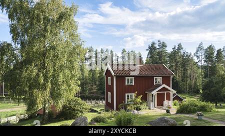 Una tipica casa svedese rossa e bianca nel piccolo e piccolo. prato verde in primo piano, piccola foresta sullo sfondo. Cielo blu con piccole nuvole. Scandinavia lan Foto Stock