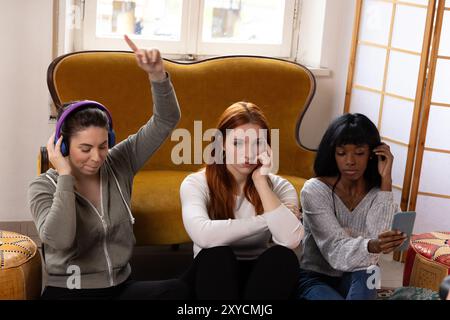Tre giovani donne sedute sul pavimento, una sta ascoltando musica con le cuffie che infastidiscono due amici annoiati Foto Stock