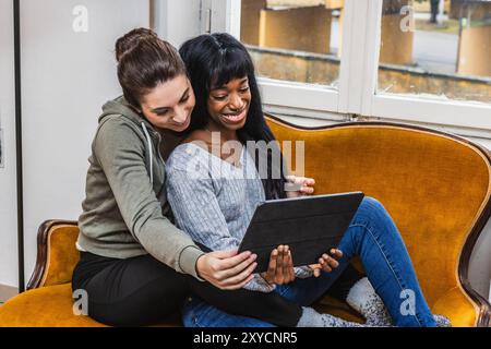 Due donne sono sedute su un divano, usano un tablet e sorridono Foto Stock
