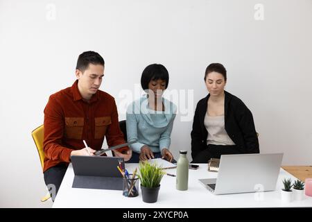 Tre colleghi mirati stanno collaborando a un progetto, utilizzando tablet e laptop digitali su una scrivania condivisa Foto Stock