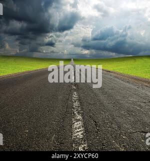 Vecchia strada di asphault rotta in un campo verde sopra il cielo blu nuvoloso Foto Stock