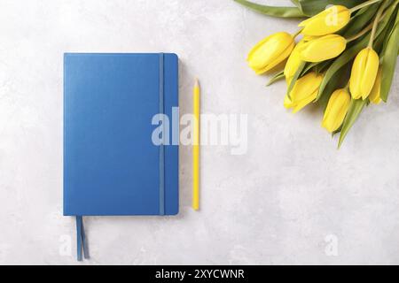 Blocco note blu, matita e bouquet di tulipani gialli primaverili su tavolo in pietra grigia, piano Foto Stock