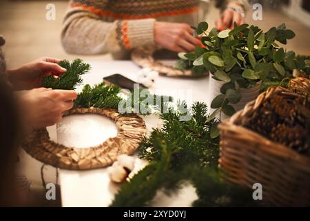 Vista ritagliata del fioraio mani rendendo ghirlanda di Natale con rami di abete e bacche decorativi sul tavolo di legno Foto Stock