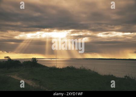 Sulla costa di Hundested. I raggi del sole si rompono attraverso il cielo drammatico attraverso le nuvole. Prato con sentiero in primo piano. Paesaggio girato in Danimarca Foto Stock